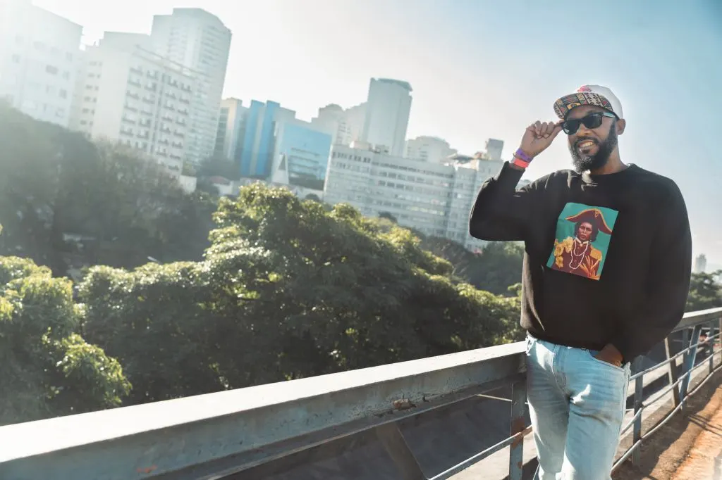 Vox Sambou on a city overpass, wearing a sweatshirt printed with the We Must Unite cover